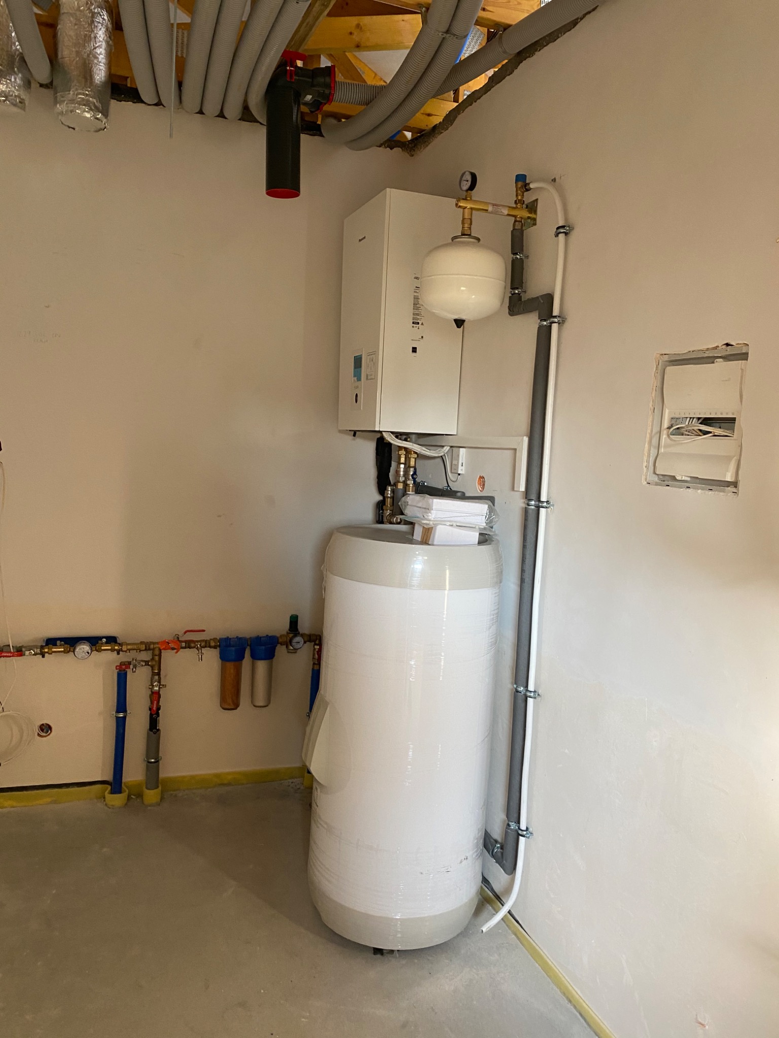 Biały, owinięty folią zasobnik ciepłej wody użytkowej, zamontowany obok białego pieca grzewczego z widocznymi rurami i zaworami, na tle surowej ściany z instalacją elektryczną w trakcie montażu.