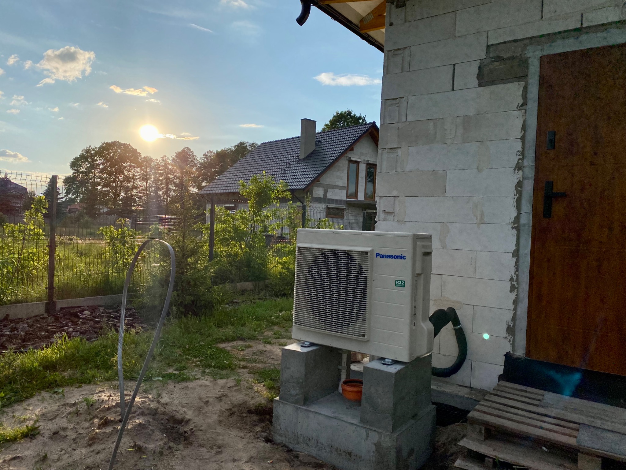 Zewnętrzna jednostka klimatyzacji Panasonic R32, zamontowana na betonowym fundamencie przy ścianie budynku z pustaków, widoczny przewód odprowadzający skropliny, w tle dom jednorodzinny...
