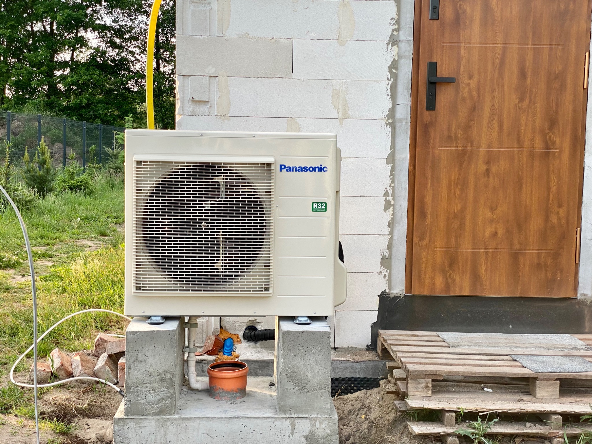 Klimatyzator Panasonic zamontowany na betonowych postumentach obok budynku z białych bloczków, widoczne elementy instalacji odprowadzającej skropliny i żółty przewód, w tle trawa i drzewa.
