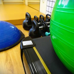 Warsztat Zdrowia - Wnętrze gabinetu z wyposażeniem do ćwiczeń: duża zielona piłka gimnastyczna, niebieska platforma BOSU, czarne kettlebells, hantle i bieżnia z żółtym akcentem.
