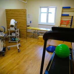 Warsztat Zdrowia - Jasne wnętrze gabinetu z drewnianą podłogą, wyposażone w bieżnię z zieloną piłką, aparat USG, stół do masażu, drabinki gimnastyczne i sprzęt rehabilitacyjny.