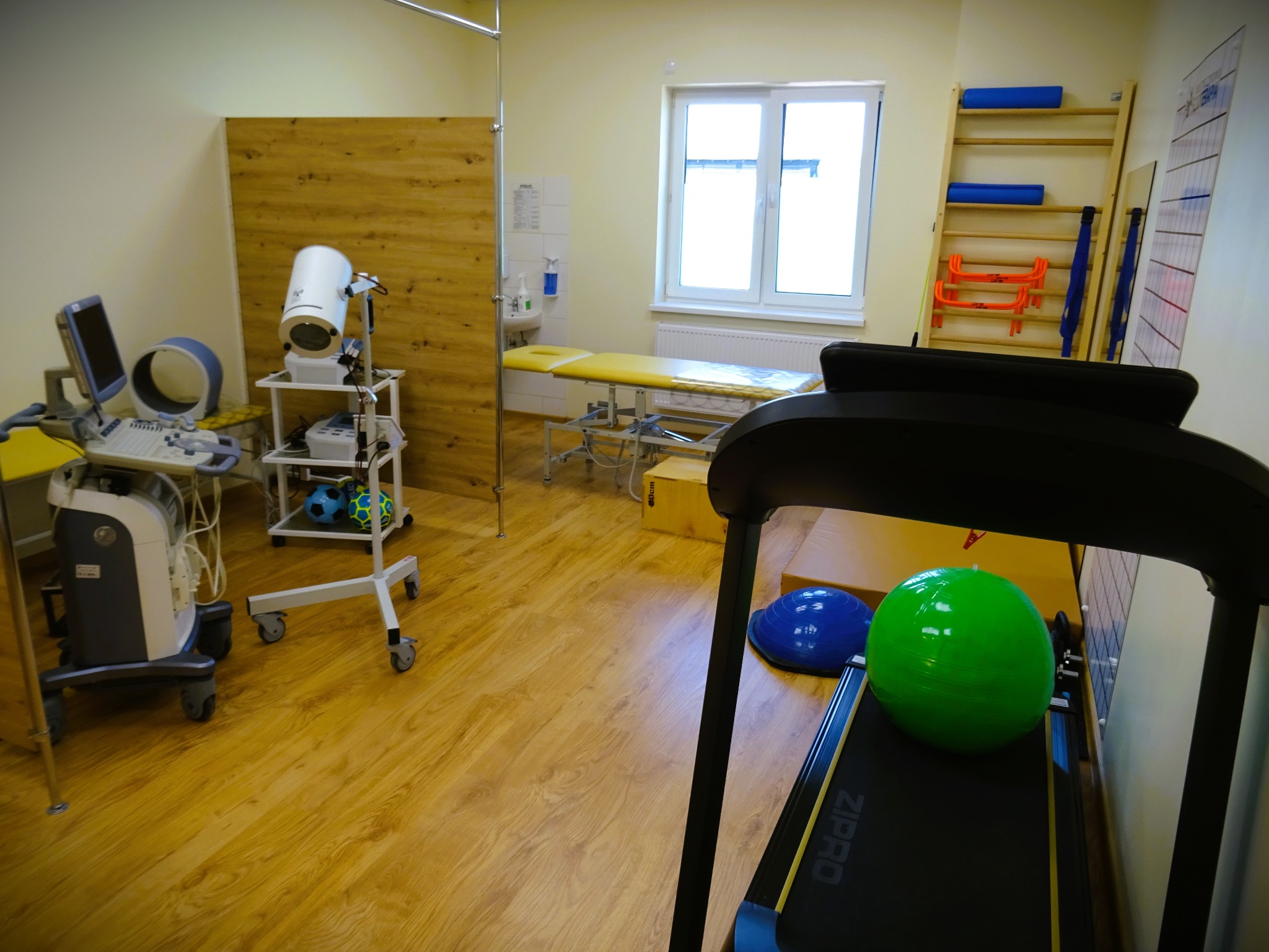 Jasne wnętrze gabinetu z drewnianą podłogą, wyposażone w bieżnię z zieloną piłką, aparat USG, stół do masażu, drabinki gimnastyczne i sprzęt rehabilitacyjny.