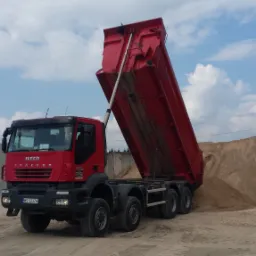 Czerwony wywrotka Iveco Trakker z podniesioną skrzynią ładunkową, wysypujący piasek na pryzmę, na tle błękitnego nieba z chmurami.