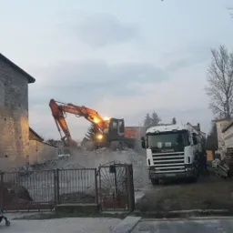 Pomarańczowa koparka Hitachi w Trzebieszowie podczas wyburzania murowanego budynku z pustaków; widoczna ciężarówka Scania, fragment ogrodzenia i spacerująca rodzina z wózkiem.