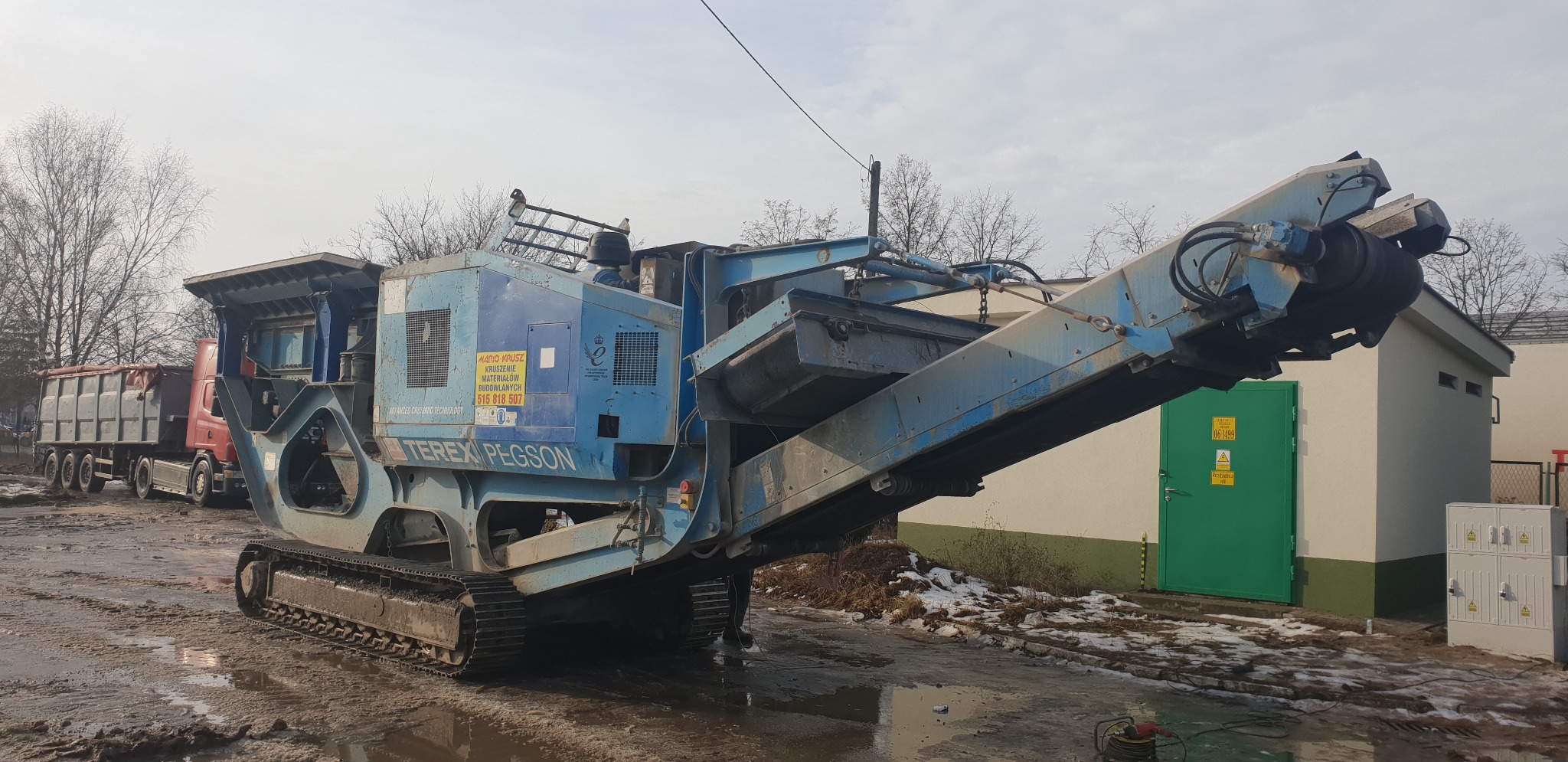 Mobilna kruszarka Terex Pegson na gąsienicach, widoczna na placu budowy z błotnistym podłożem i zaparkowaną ciężarówką w tle. Maszyna w kolorze niebieskim, z żółtą naklejką informacyjną...