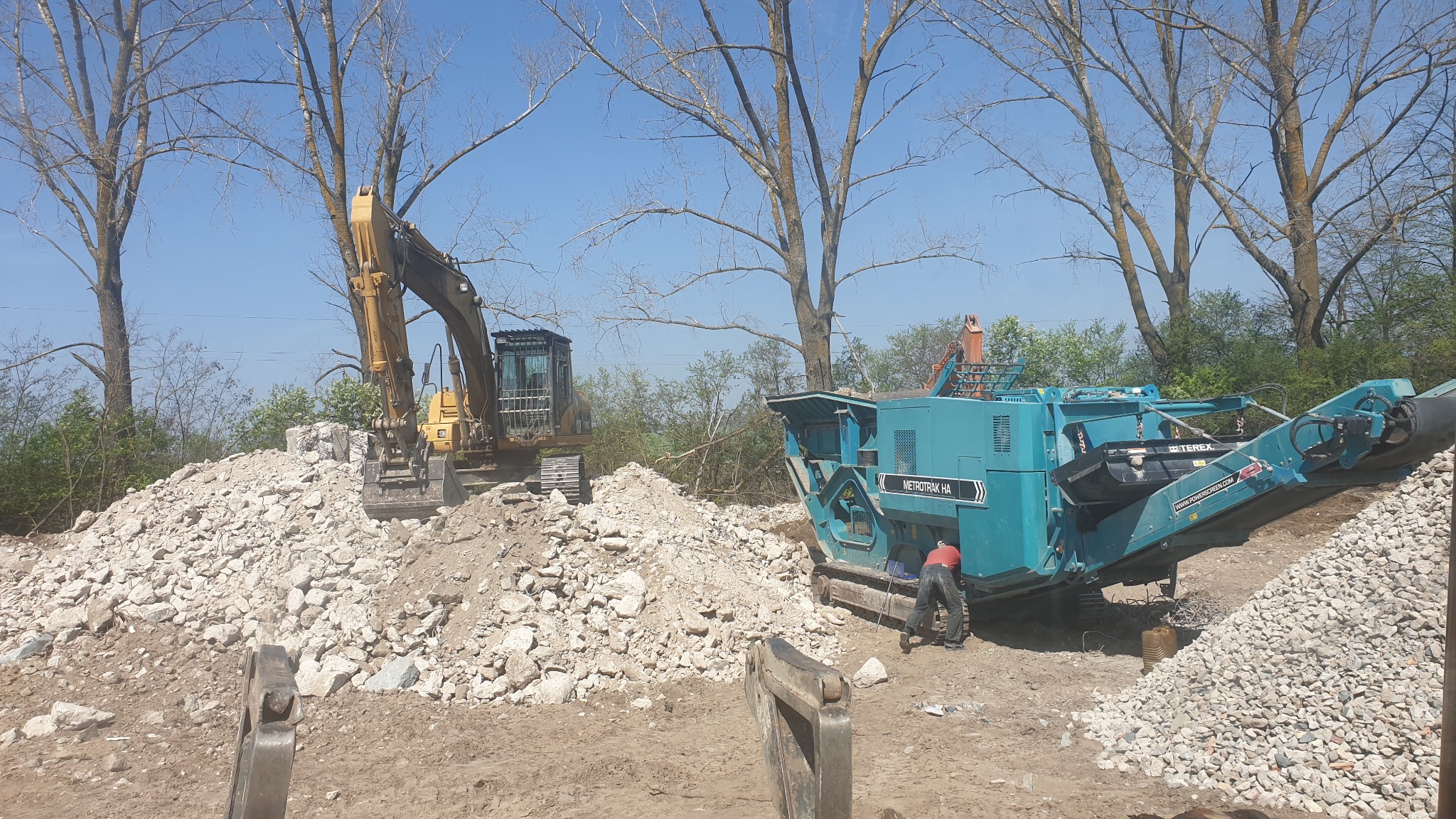 Żółta koparka i mobilny kruszarz do gruzu Metrotrak HA na placu budowy, operator w czerwonej koszuli przy maszynie, hałda gruzu w tle.
