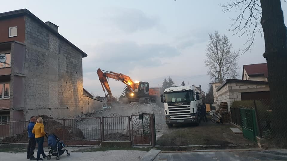 Pomarańczowa koparka Hitachi w Trzebieszowie podczas wyburzania murowanego budynku z pustaków; widoczna ciężarówka Scania, fragment ogrodzenia i spacerująca rodzina z wózkiem.
