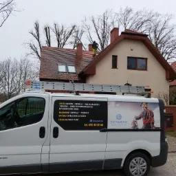 Pracownik w żółtym kombinezonie na dachu z czerwonej dachówki, widoczny zza białego samochodu dostawczego z reklamą firmy Skywork24 oferującej usługi alpinistyczne, mycie dachów i elewacji.