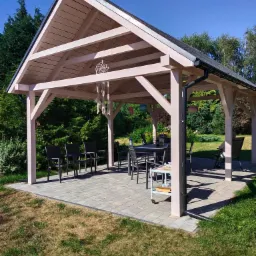 Jasna, drewniana altana z szarym dachem i rynnami, ustawiona na kostce brukowej w ogrodzie, z widocznym stołem, krzesłami i łapaczem snów.