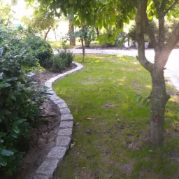 Krawężnik z szarej kostki granitowej oddzielający rabatę z zielonymi krzewami od trawnika w słonecznym ogrodzie.