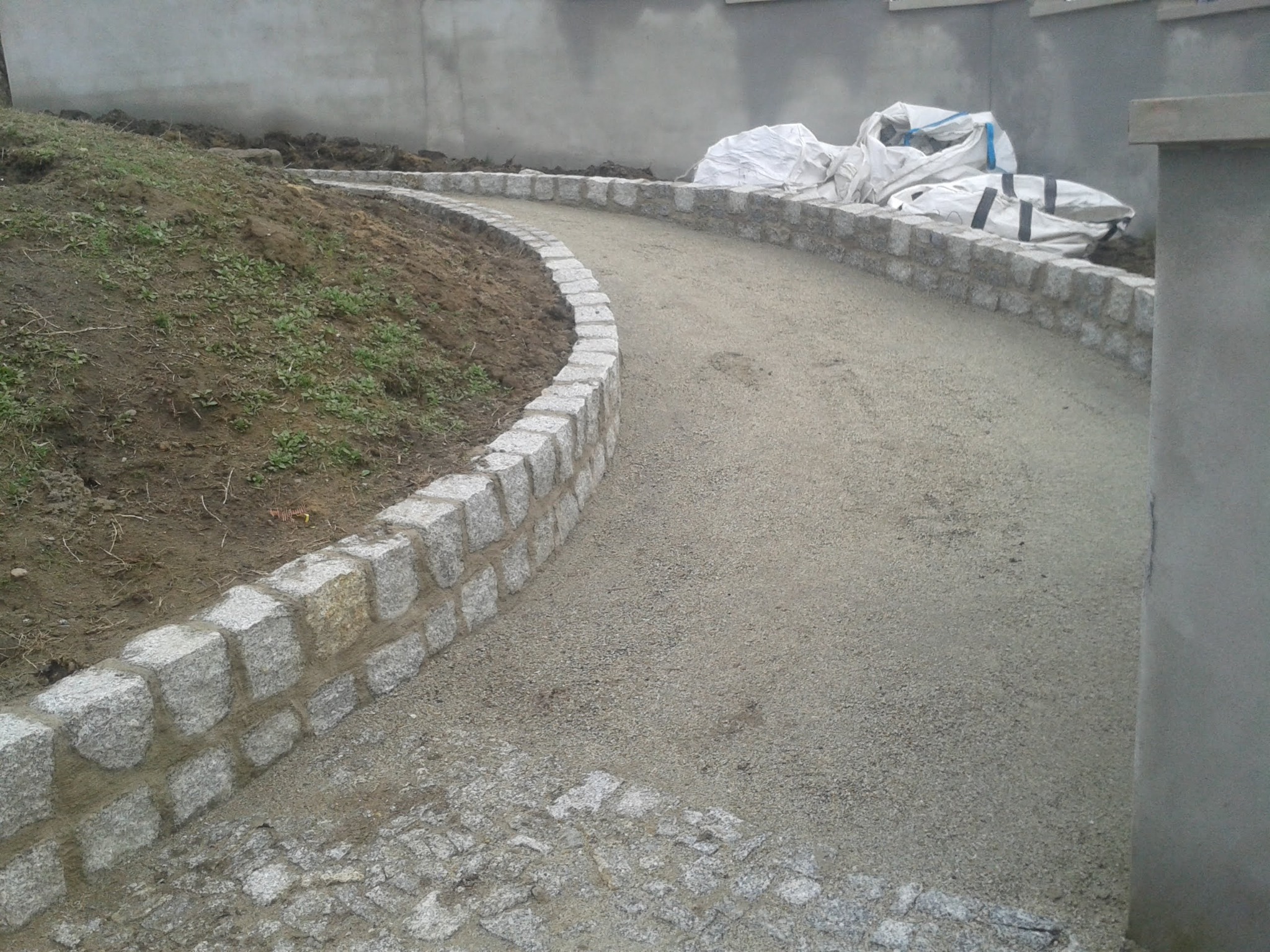 Ścieżka z drobnego kruszywa ograniczona obrzeżem z kostki granitowej, z workami materiałów budowlanych w tle, na etapie realizacji.