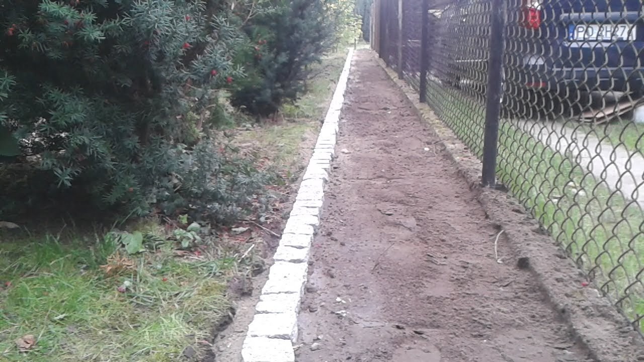Krawężnik z jasnych kostek oddzielający ścieżkę z ubitej ziemi od zieleni, widok na ogrodzenie z siatki i zaparkowany samochód w tle.