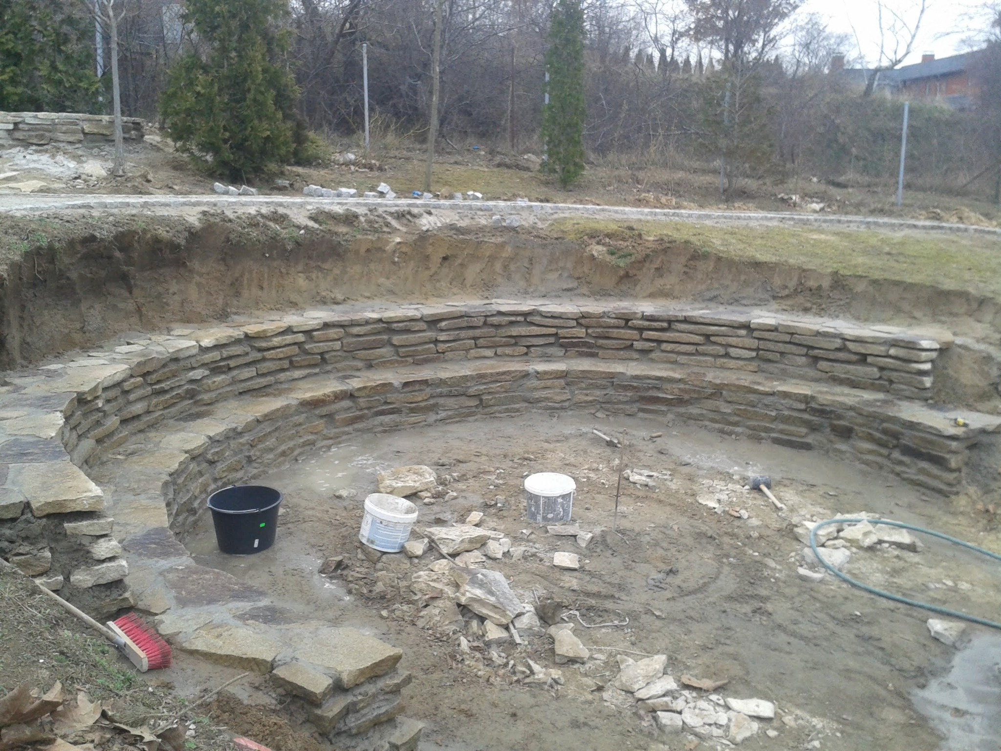 Półokrągła konstrukcja z kamienia w trakcie budowy, widoczne narzędzia, wiaderka i gruz na placu budowy, otoczenie ziemne z drzewami w tle.