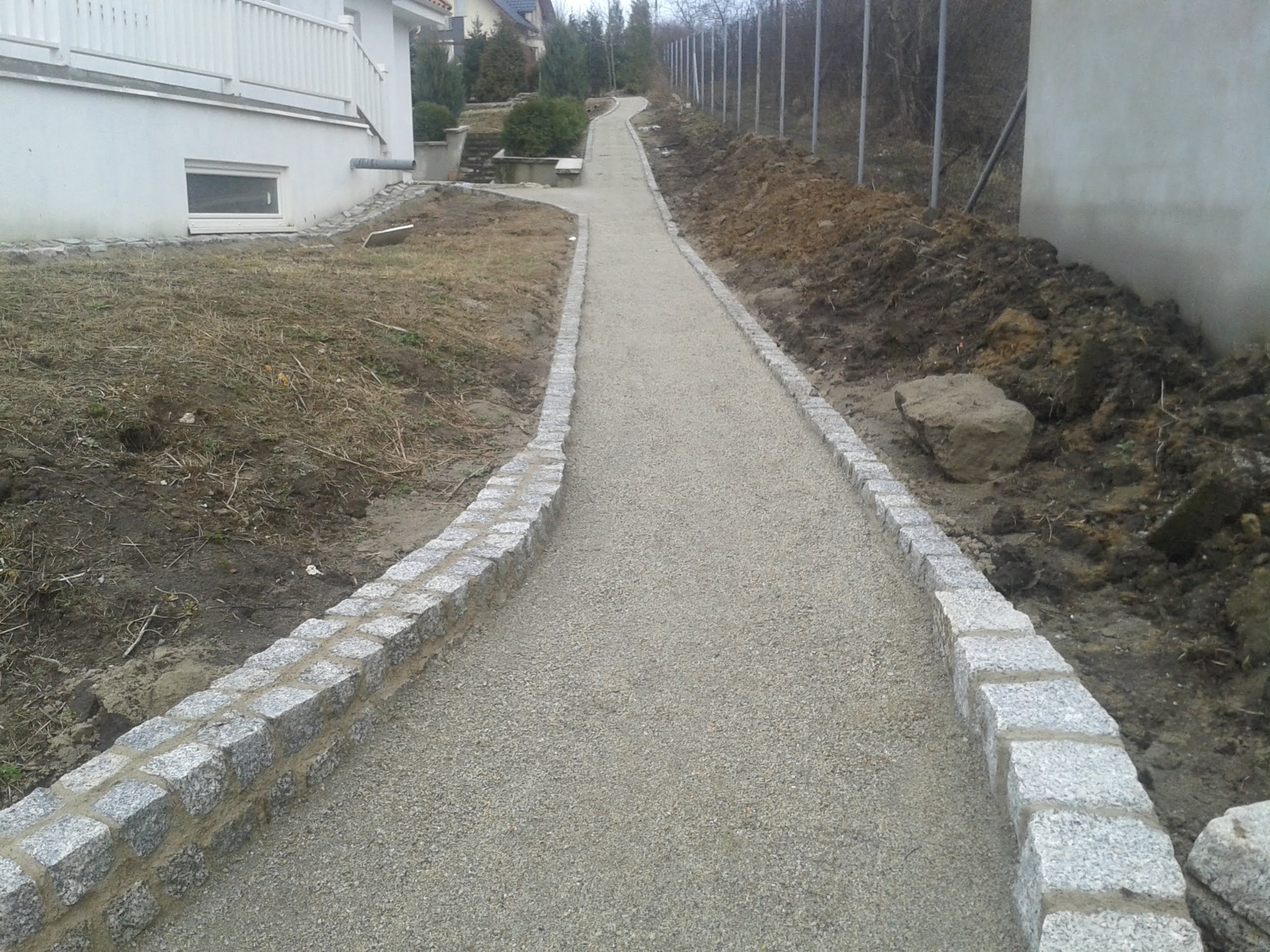 Ścieżka z szarego kruszywa ograniczona kostką granitową, w trakcie budowy, widok na dom i ogrodzenie.