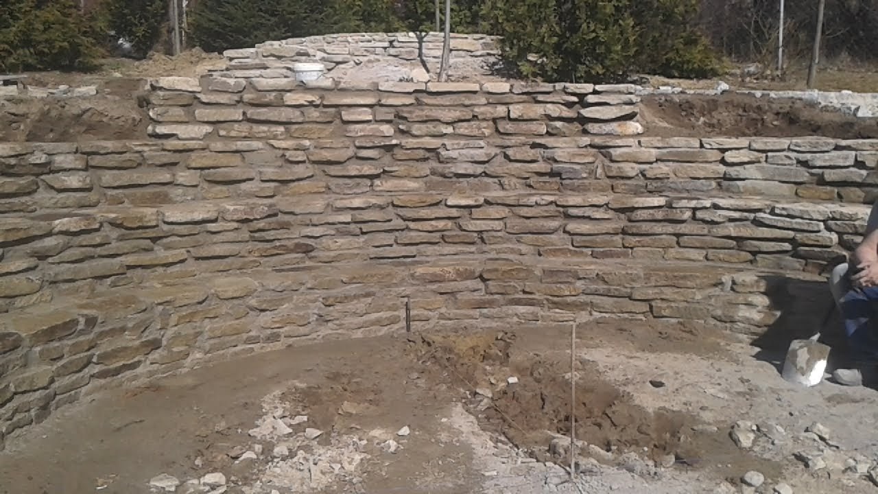 Budowa kaskadowego muru oporowego z kamienia naturalnego w ogrodzie, widoczne warstwy kamieni i przygotowane podłoże.