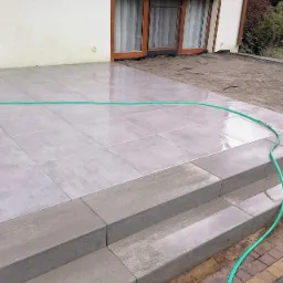 Szare płytki tarasowe ułożone na zewnątrz domu, z trzema schodkami z tego samego materiału, zielony wąż ogrodowy leży na powierzchni, w tle okno z brązowymi roletami.