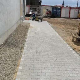 Układanie szarej kostki brukowej typu I na nowym chodniku, widoczny pracownik zagęszczający powierzchnię, obok budynek i kontenery budowlane.