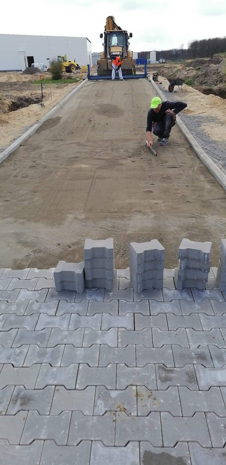 Układanie szarej kostki brukowej typu 'H' na przygotowanym podłożu; w tle pracownicy i ciężki sprzęt budowlany na placu budowy.