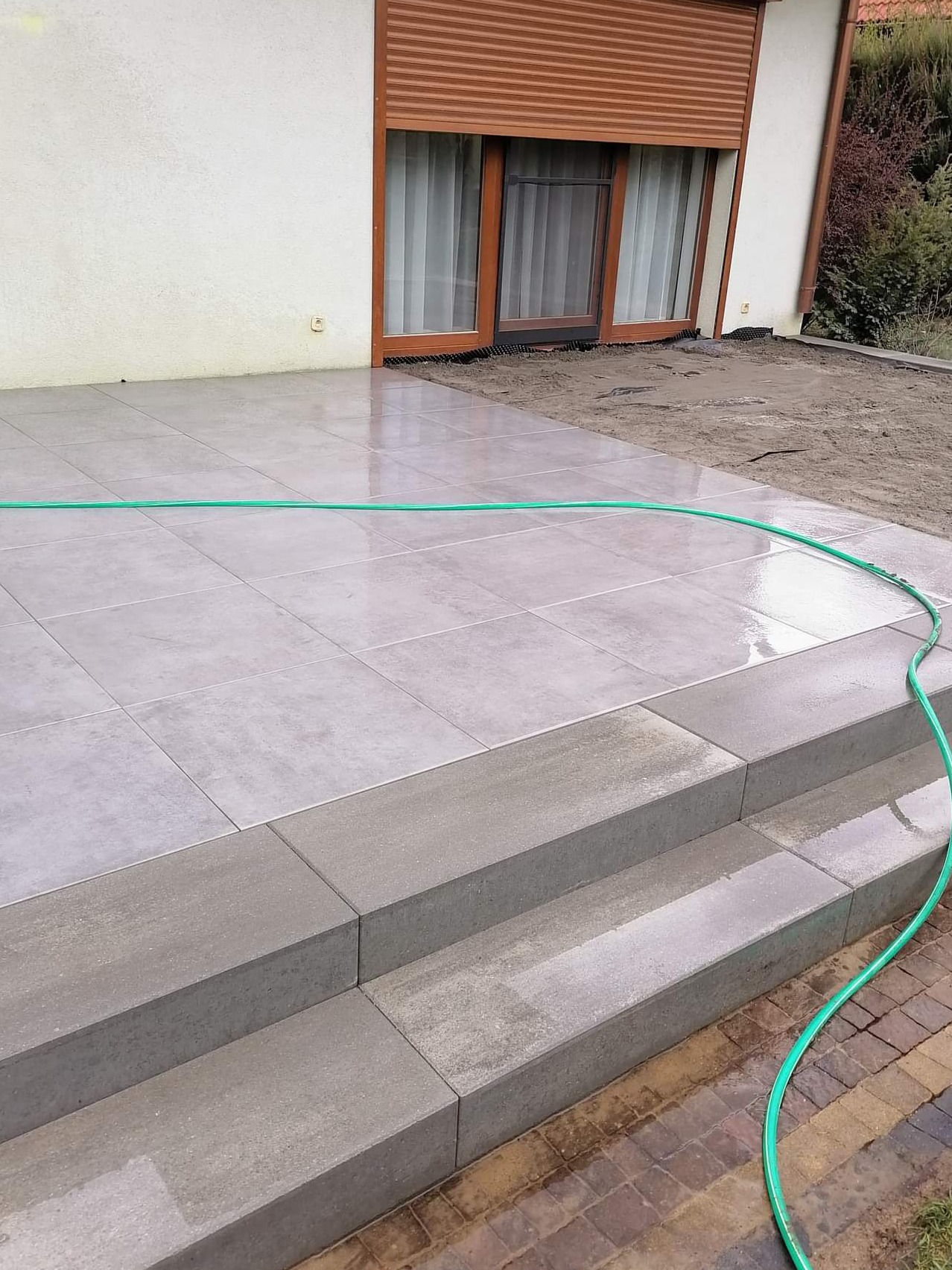 Szare płytki tarasowe ułożone na zewnątrz domu, z trzema schodkami z tego samego materiału, zielony wąż ogrodowy leży na powierzchni, w tle okno z brązowymi roletami.