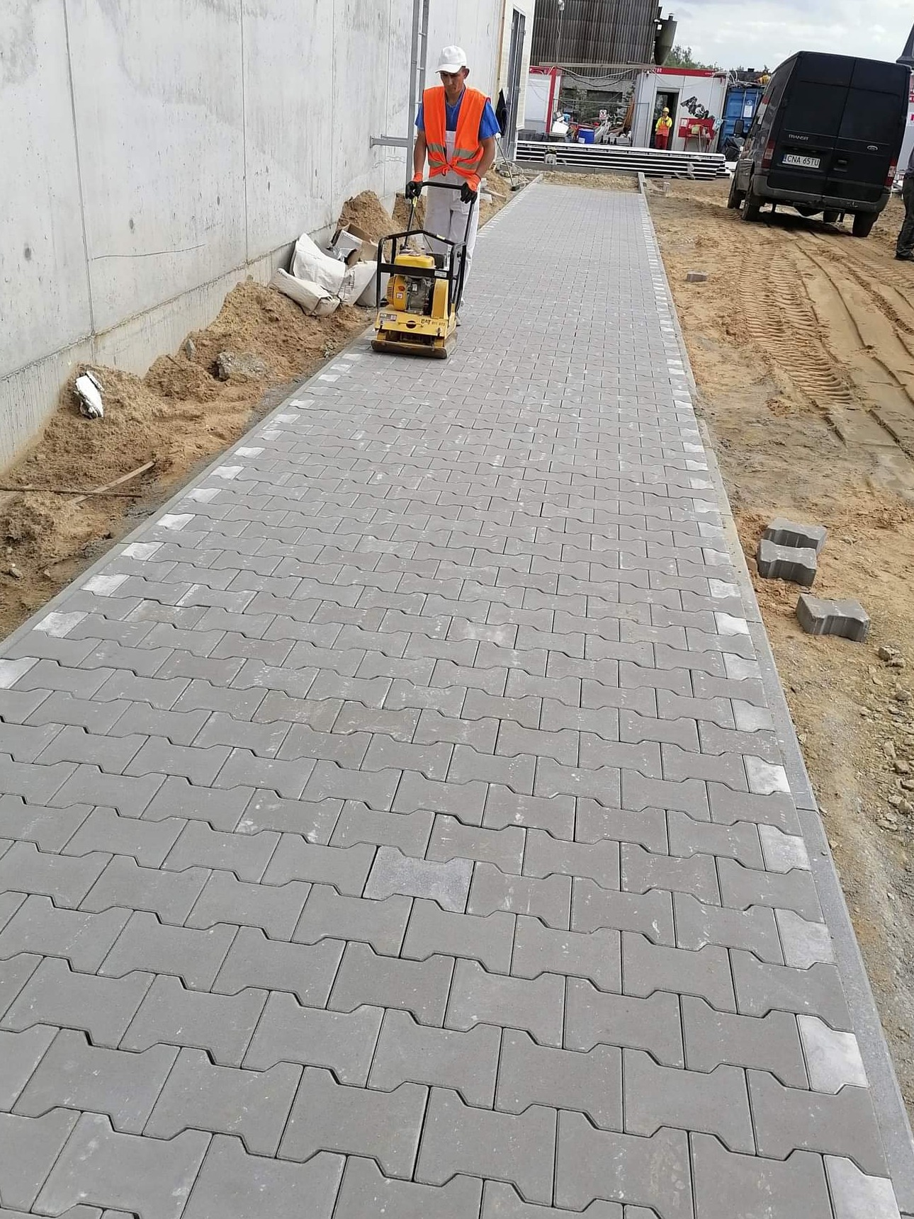 Pracownik w kamizelce odblaskowej zagęszcza nowo ułożoną, szarą kostkę betonową typu 'H' wzdłuż betonowego muru, z zapasem kostki obok i zaparkowanym czarnym vanem w tle.
