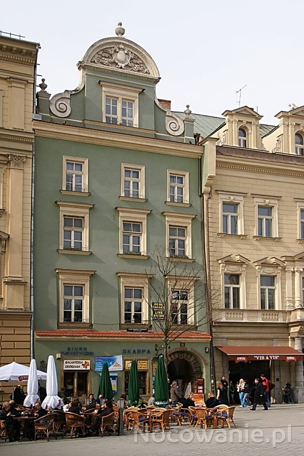 Elewacja kamienicy z szyldem 'Biuro Turystyczne', stoliki kawiarniane na pierwszym planie, widok z zewnątrz.