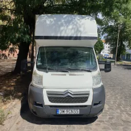 Biały samochód dostawczy Citroen z wysoką zabudową, zaparkowany na brukowanej ulicy w cieniu drzewa, widok z przodu.