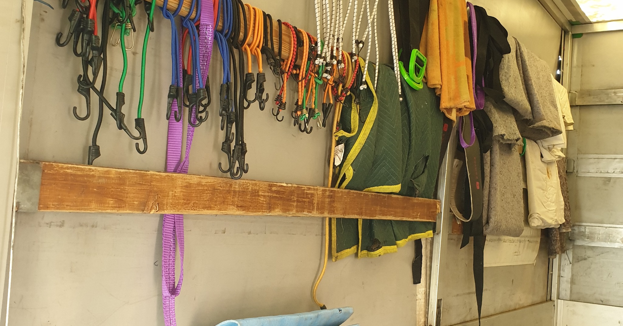 Wnętrze furgonetki: gumowe linki z haczykami w różnych kolorach zawieszone na drewnianej listwie, obok koce transportowe i zabezpieczenia do mebli.