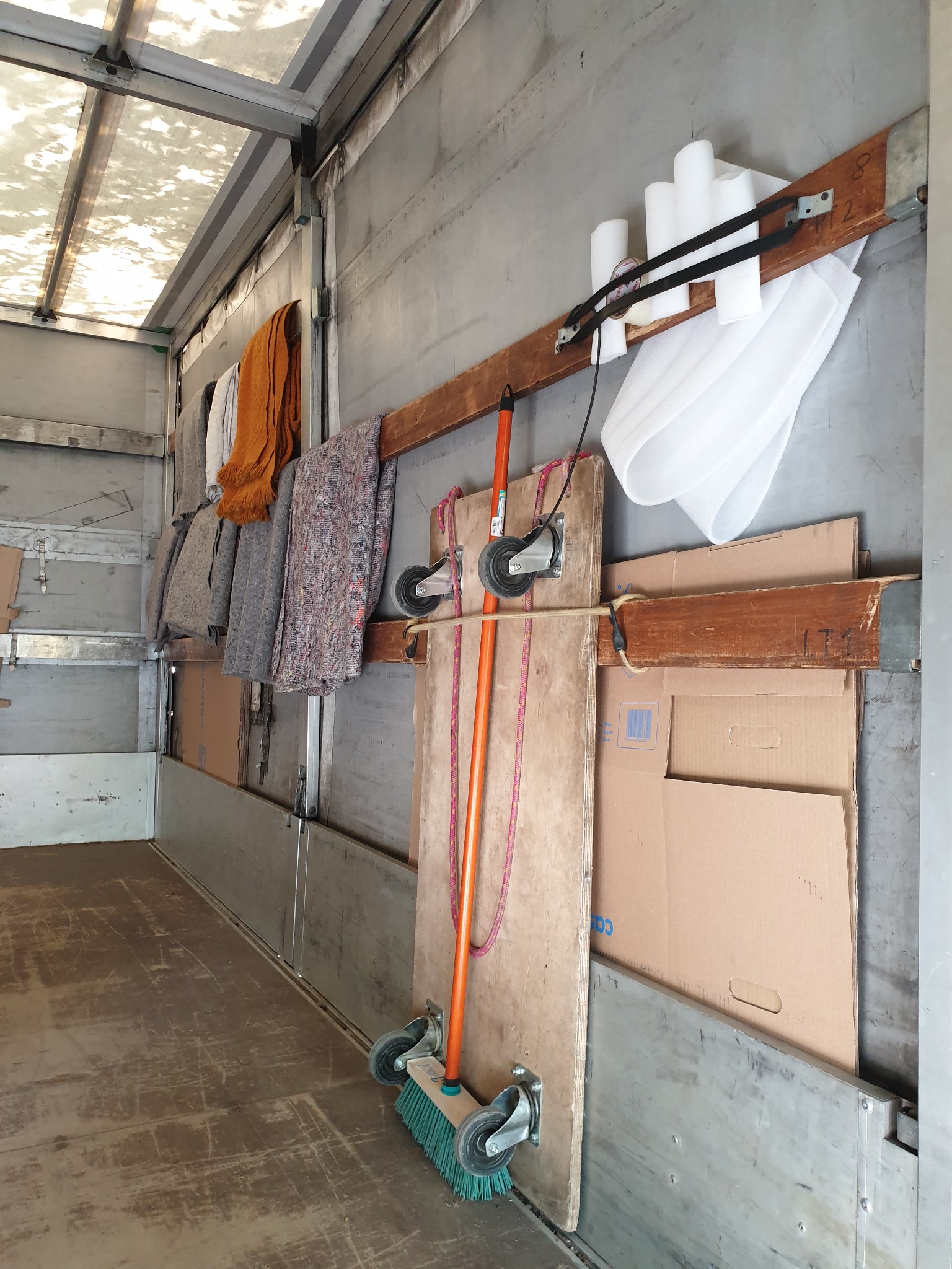 Wnętrze paki samochodu dostawczego, widoczne koce transportowe, kartony, rolki folii zabezpieczającej i szczotka do sprzątania, przymocowane do ścian i drewnianych belek.