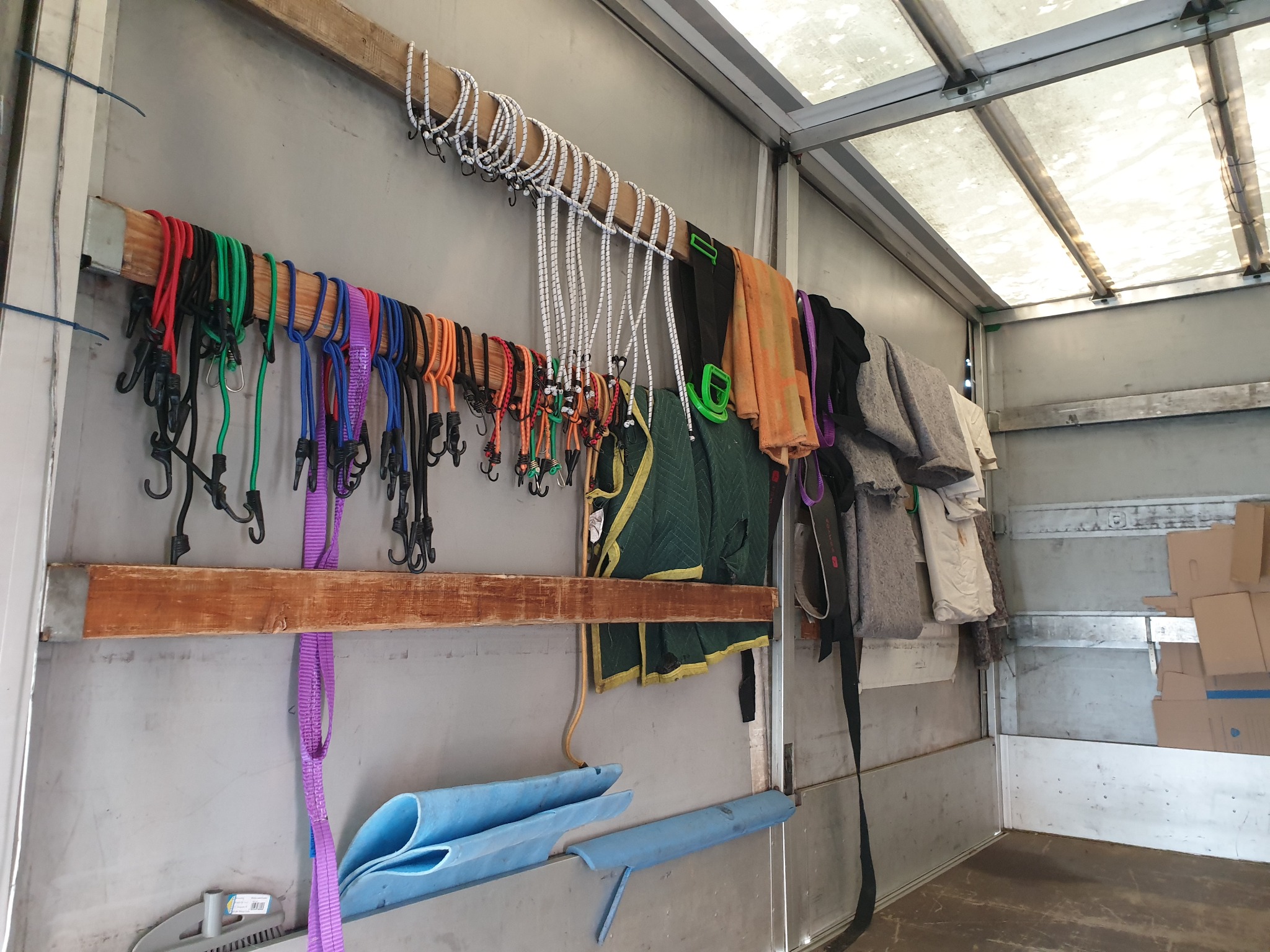 Wnętrze furgonetki przeprowadzkowej z zabezpieczeniami: kolorowe gumy mocujące z hakami, białe linki elastyczne, koce transportowe i niebieskie osłony rur, przymocowane do drewnianych listew...