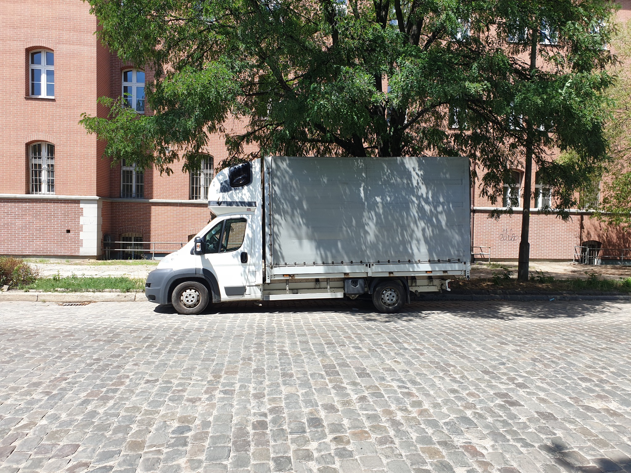 Biała furgonetka z plandeką zaparkowana na brukowanej ulicy, w tle ceglany budynek i drzewo w słoneczny dzień.