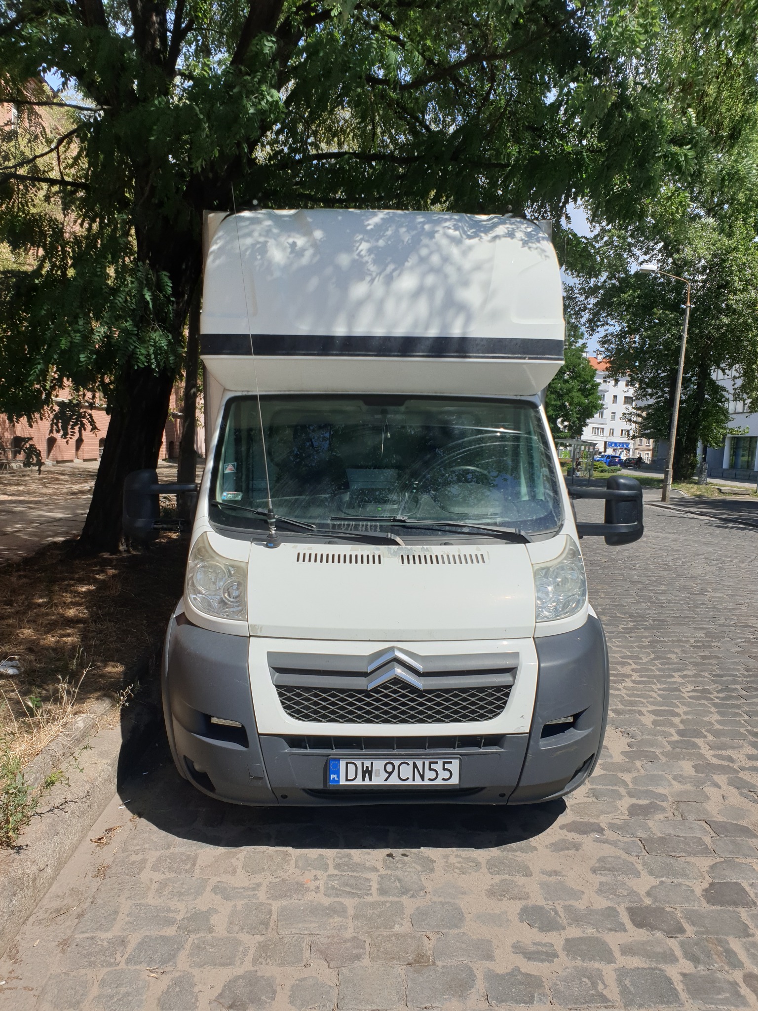 Biały samochód dostawczy Citroen z wysoką zabudową, zaparkowany na brukowanej ulicy w cieniu drzewa, widok z przodu.