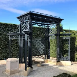 Ozdobna, metalowa altana ogrodowa w kolorze grafitowym, z ażurowymi ścianami i kamiennym stolikiem wewnątrz, na tle zielonego żywopłotu i błękitnego nieba.