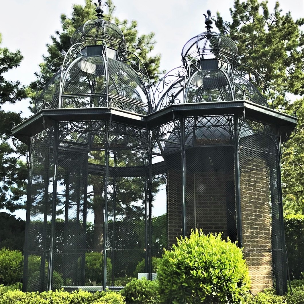 Dwie połączone, ozdobne, metalowe altany z siatki, zwieńczone kopułami, otoczone zielenią ogrodową i drzewami.