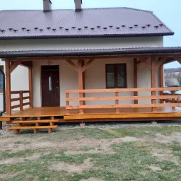 Zadaszony drewniany taras przed domem z brązowym dachem, widoczne trzy stopnie wejściowe i drewniana balustrada, teren wokół w trakcie prac wykończeniowych.