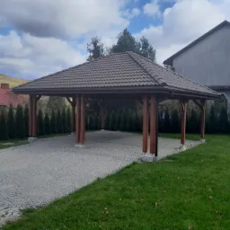 Zadaszona konstrukcja z brązową dachówką i drewnianymi słupami na żwirowym podłożu, otoczona tujami i budynkami w tle, widok z zewnątrz.