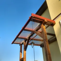 Drewniana konstrukcja z przezroczystym zadaszeniem i wiszącą lampą łańcuchową, przylegająca do ściany budynku, widok z dołu na tle błękitnego nieba.