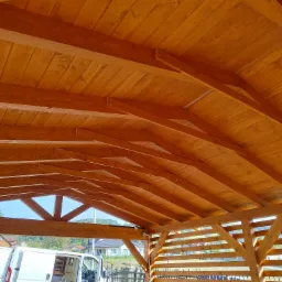 Drewniana konstrukcja z widocznym dachem z desek i częściowo osłoniętymi bokami, w tle brama i samochód dostawczy.