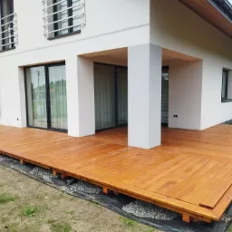 Nowoczesny taras drewniany z desek w odcieniach brązu, przylegający do białego domu z dużymi oknami. Widoczne kamienie pod konstrukcją tarasu.