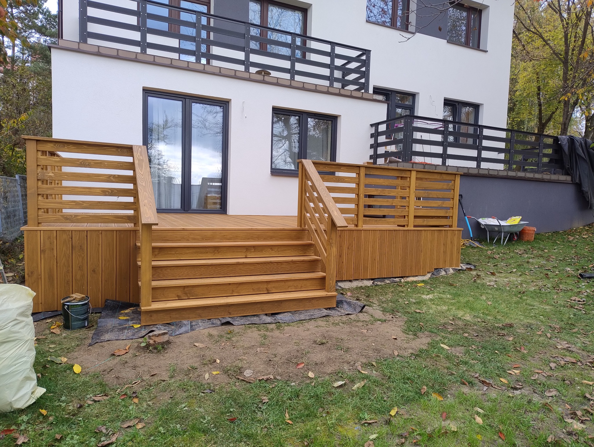 Drewniany taras z ażurową balustradą i schodami prowadzącymi do domu. Widoczne narzędzia i materiały budowlane sugerują etap końcowy prac.