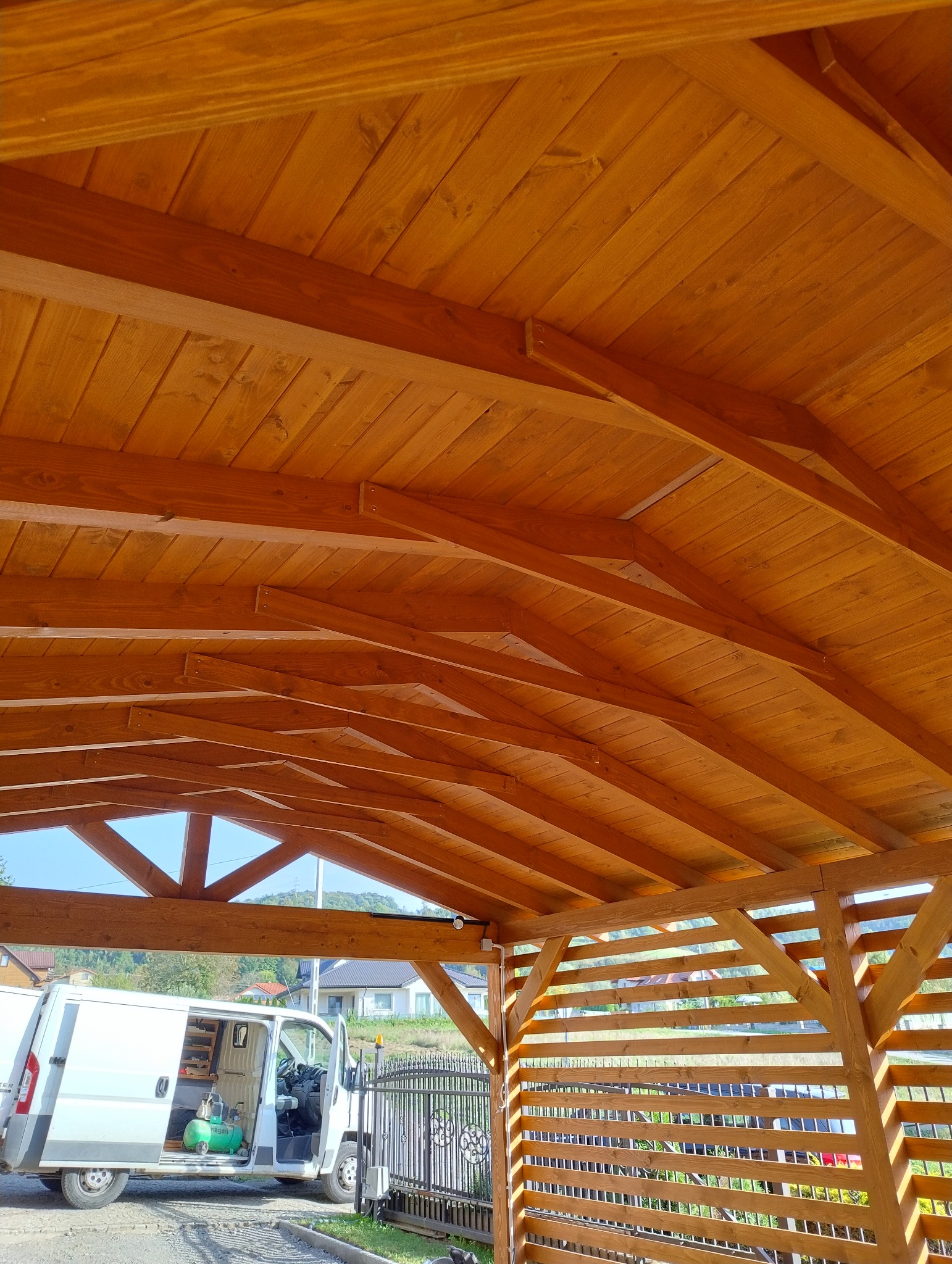 Drewniana konstrukcja z widocznym dachem z desek i częściowo osłoniętymi bokami, w tle brama i samochód dostawczy.