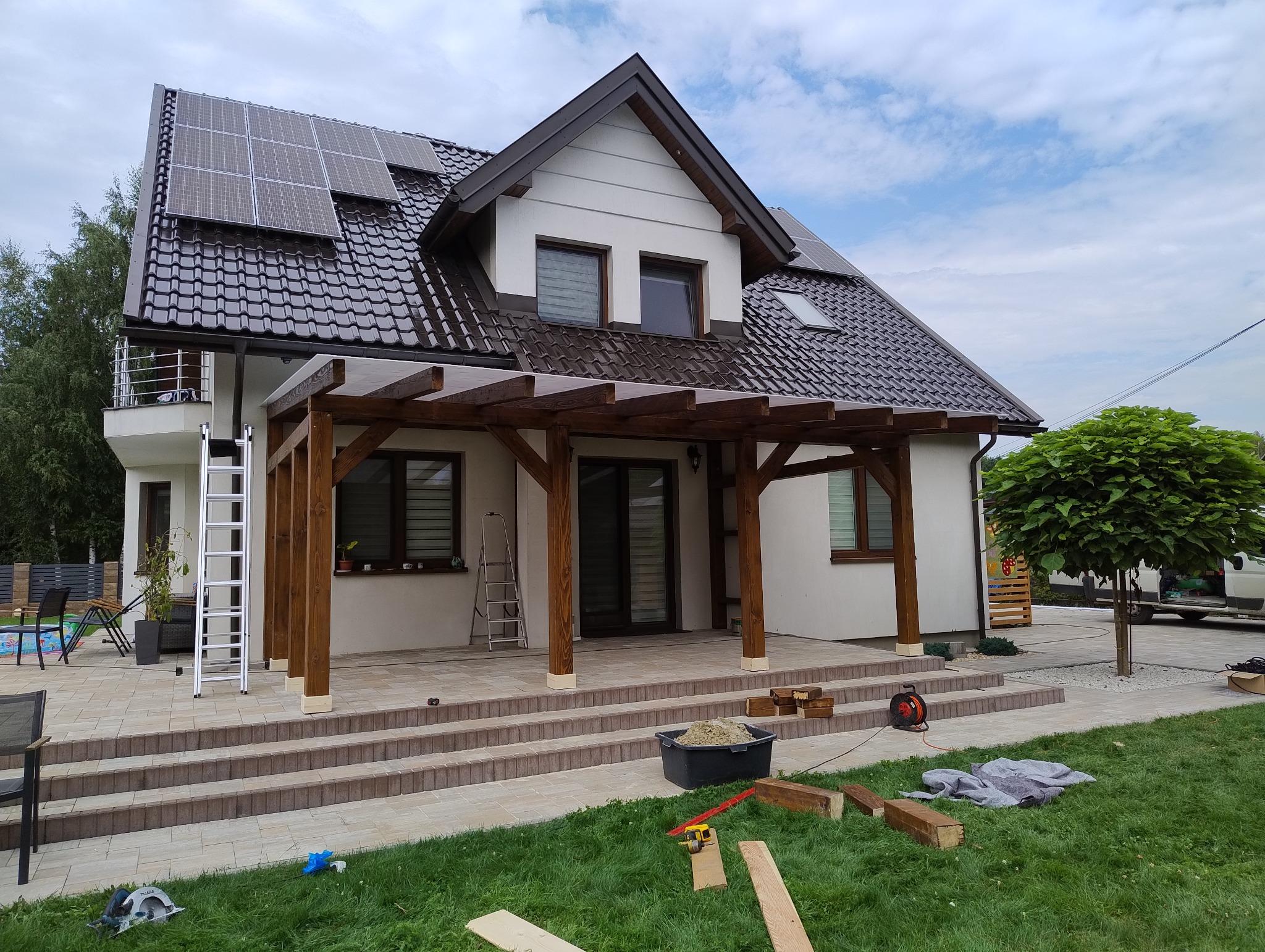 Montaż drewnianej pergoli przy domu z panelami słonecznymi. Widoczne narzędzia, schody z kostki, trawnik. Pergola z brązowych belek, częściowo zadaszona.