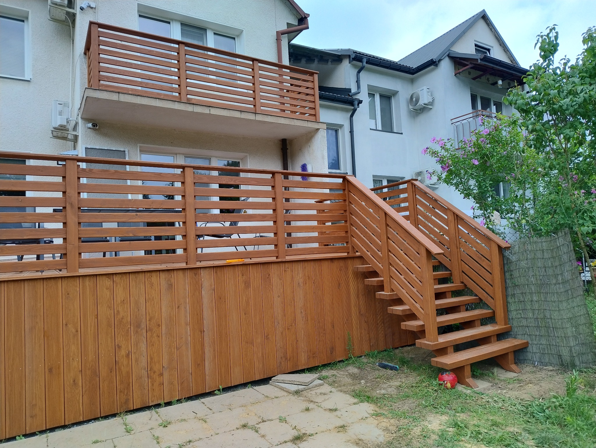 Nowoczesny, drewniany taras z balustradą i schodami, przylegający do budynku. Widoczne poziome deski balustrady i pionowe deski obudowy tarasu. Schody z małą ławeczką.