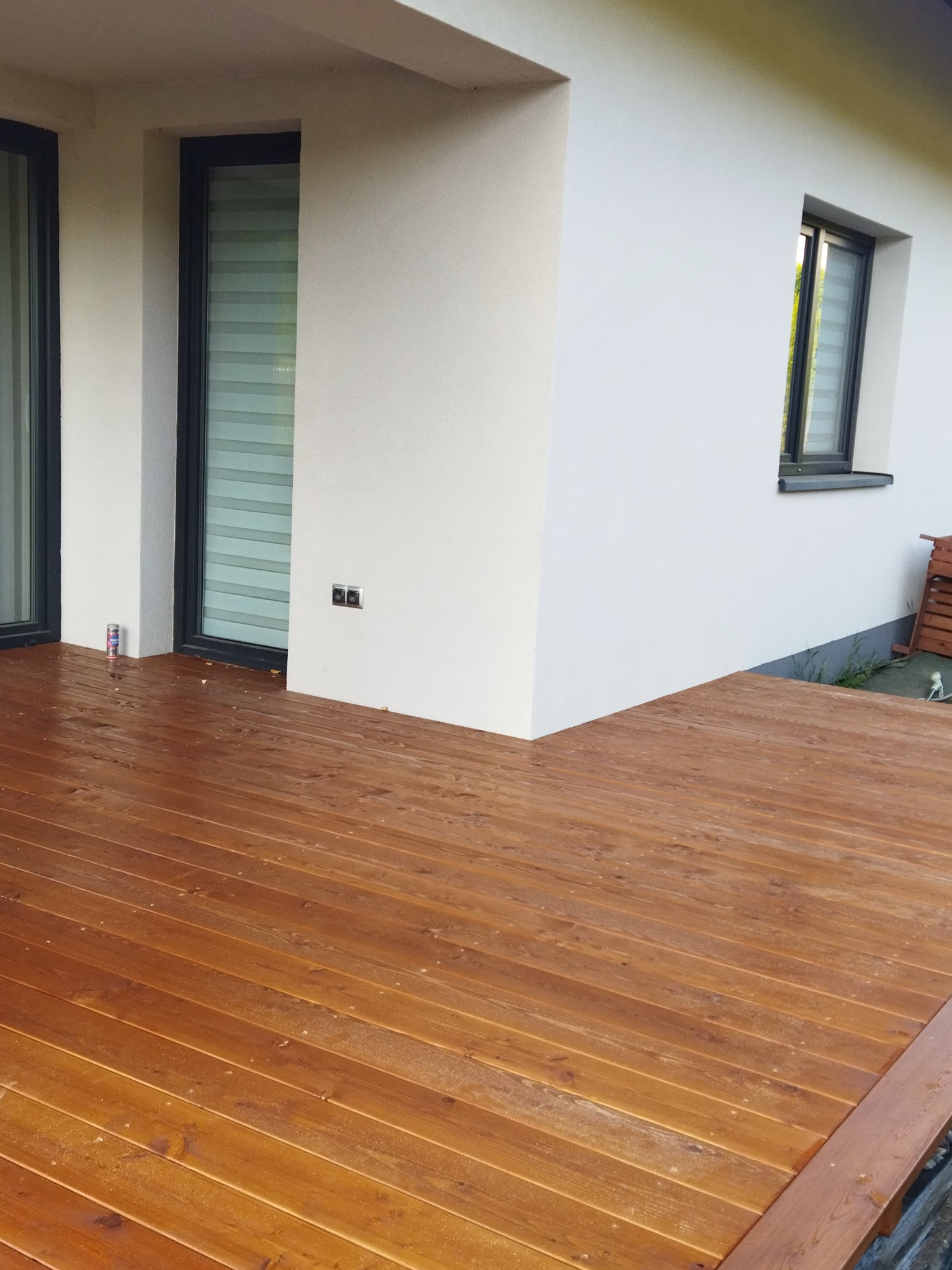 Świeżo polakierowany taras drewniany z widoczną strukturą drewna, przylegający do białego budynku z oknami. Na tarasie leży pył po szlifowaniu.