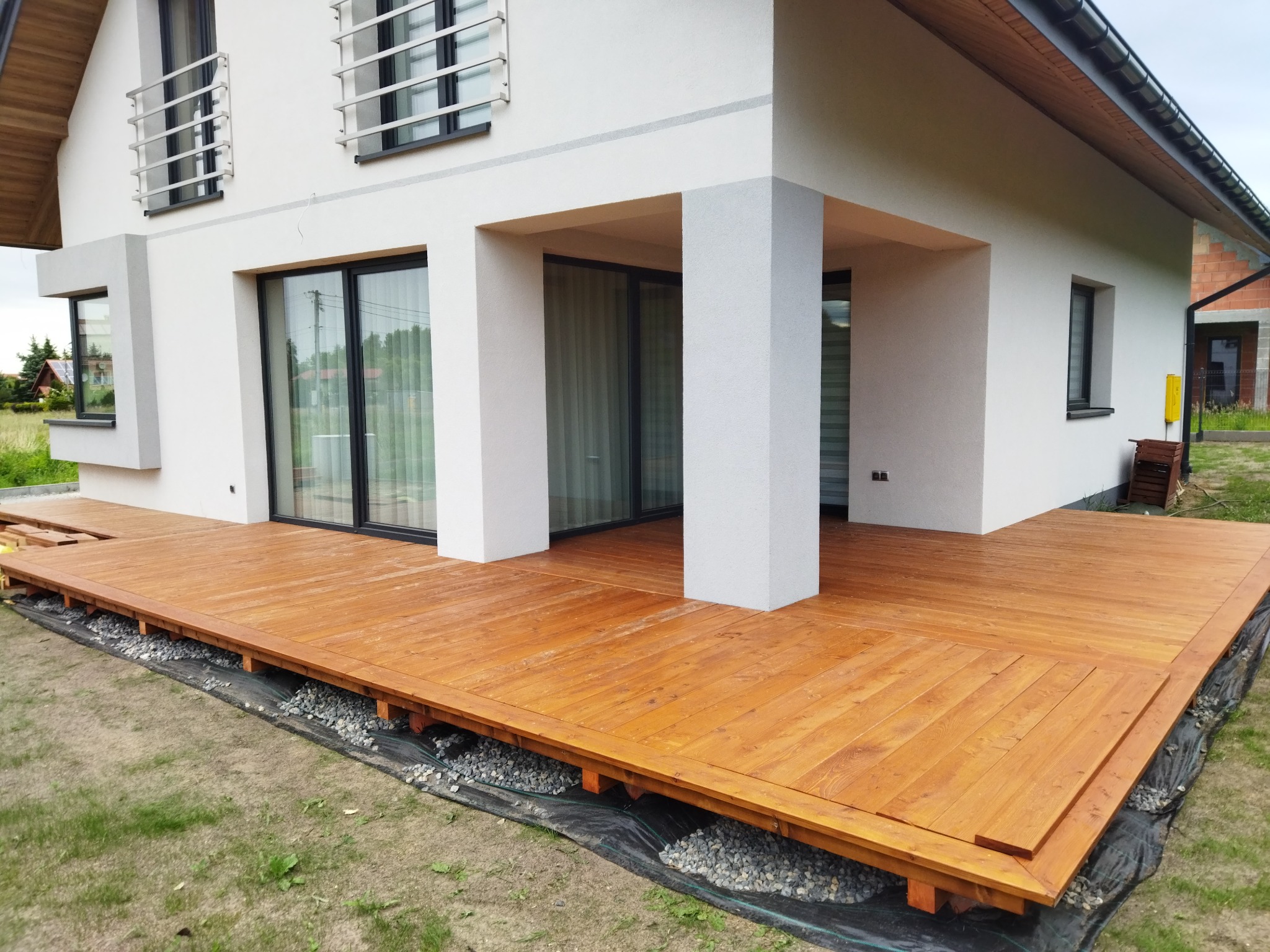 Nowoczesny taras drewniany z desek w odcieniach brązu, przylegający do białego domu z dużymi oknami. Widoczne kamienie pod konstrukcją tarasu.