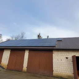 Instalacja paneli słonecznych na dachu budynku gospodarczego z trzema bramami garażowymi, widok z dołu na pochmurne niebo.