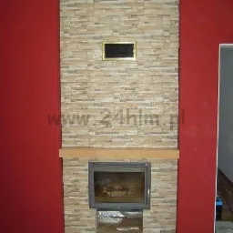 Wykończony kominek z obudową z kamienia dekoracyjnego i metalowym wkładem, czerwone ściany po bokach, brązowa podłoga z płytek.