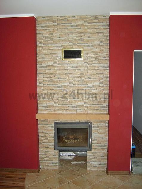 Wykończony kominek z obudową z kamienia dekoracyjnego i metalowym wkładem, czerwone ściany po bokach, brązowa podłoga z płytek.