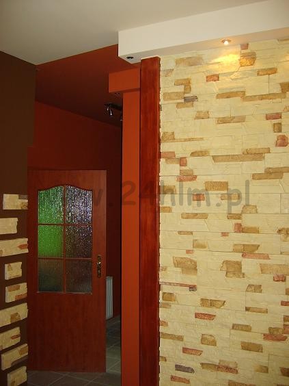 Dekoracyjna ściana z kamienia elewacyjnego w odcieniach beżu i brązu, sąsiadująca z pomarańczową ścianą i drewnianą ościeżnicą drzwi z szybą.