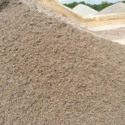 Duża pryzma piasku kwarcowego w odcieniach beżu i brązu, z widocznymi ziarnami, na tle innych pryzm piasku o różnych kolorach i zieleni drzew w oddali.