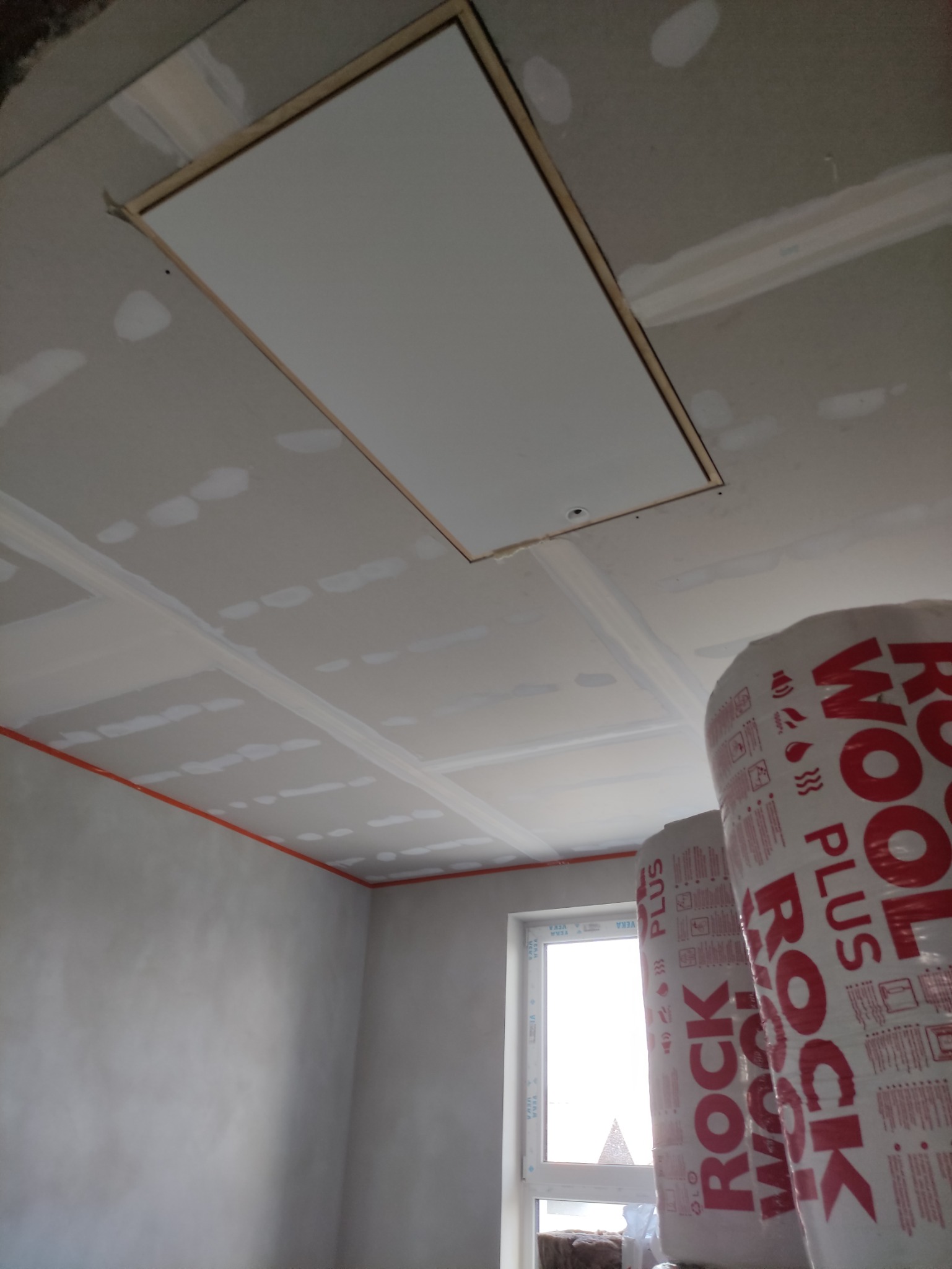 Montaż sufitu podwieszanego z płyty gipsowo-kartonowej, widoczne łączenia szpachlowane, otwór rewizyjny z klapą w suficie oraz rolki wełny mineralnej Rockwool w pomieszczeniu z oknem.